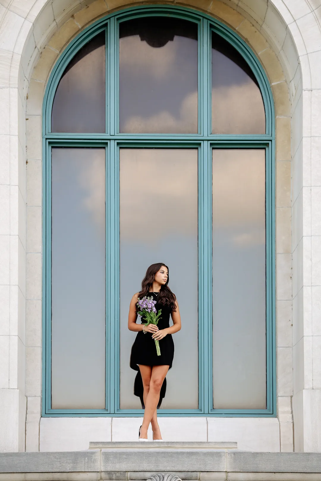 Senior in a black dress holding lavender flowers, framed by a grand arched window