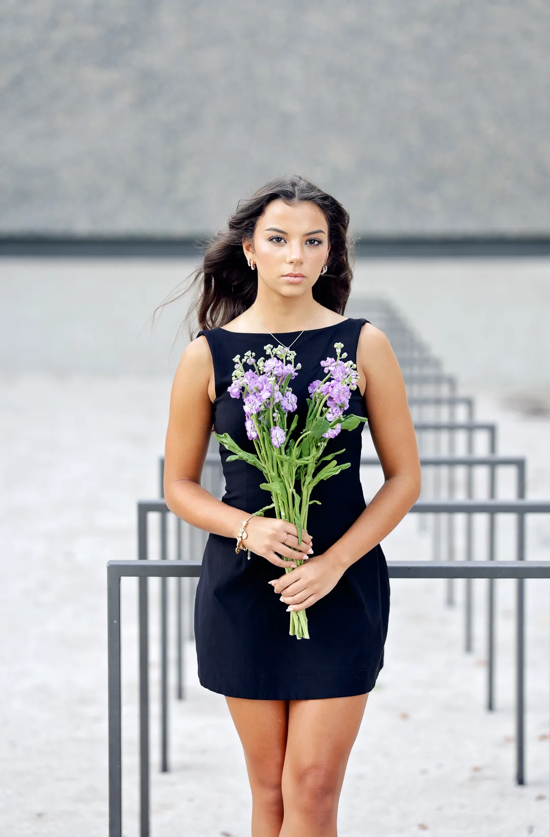 Senior in a black dress holding lavender stock flowers among steel railings