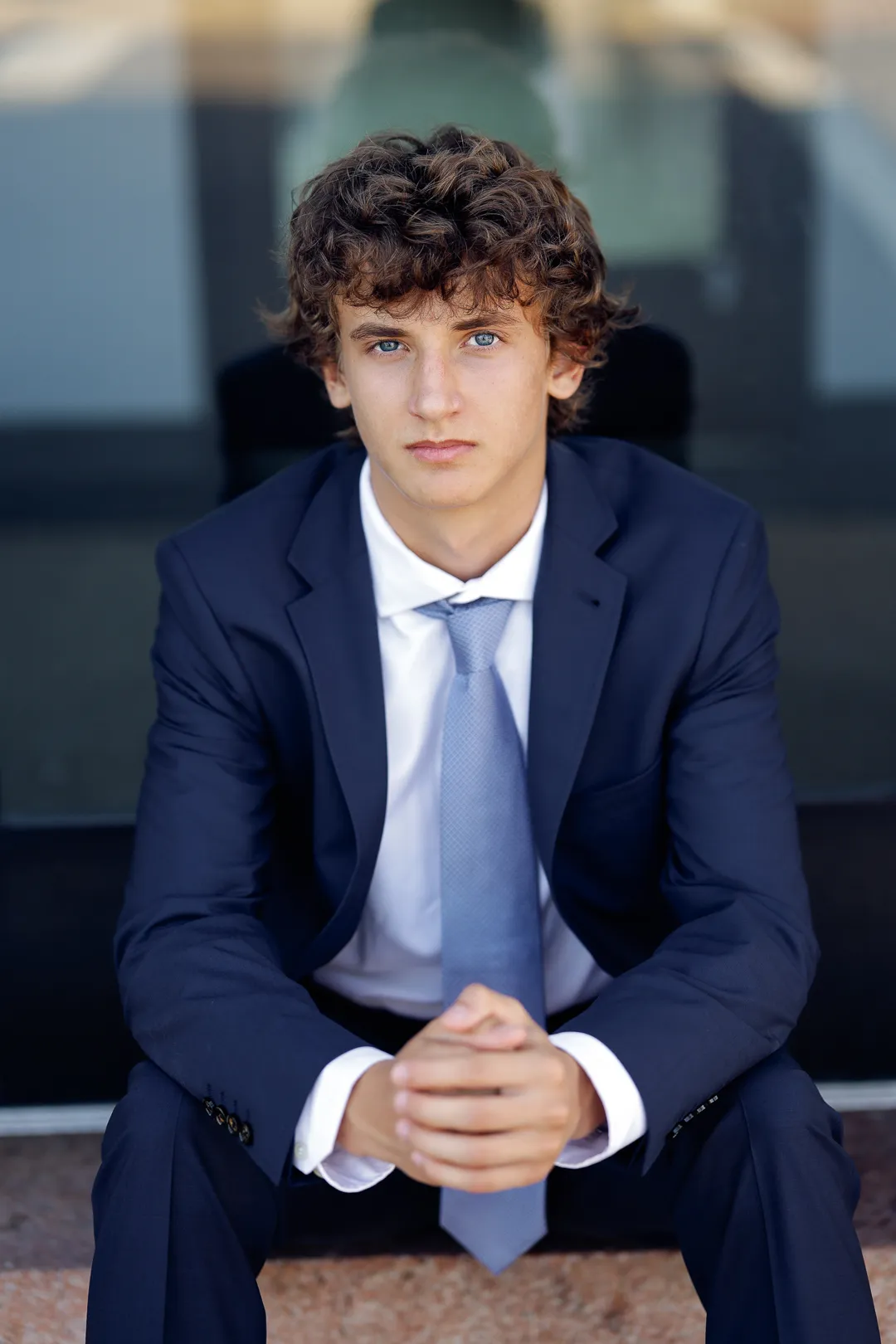 Senior in a navy suit and blue tie seated with hands clasped