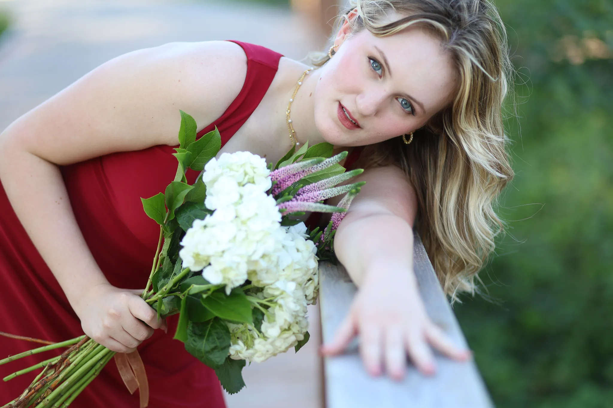 Senior in a red dress holding a hydrangea bouquet, leaning on a bridge rail