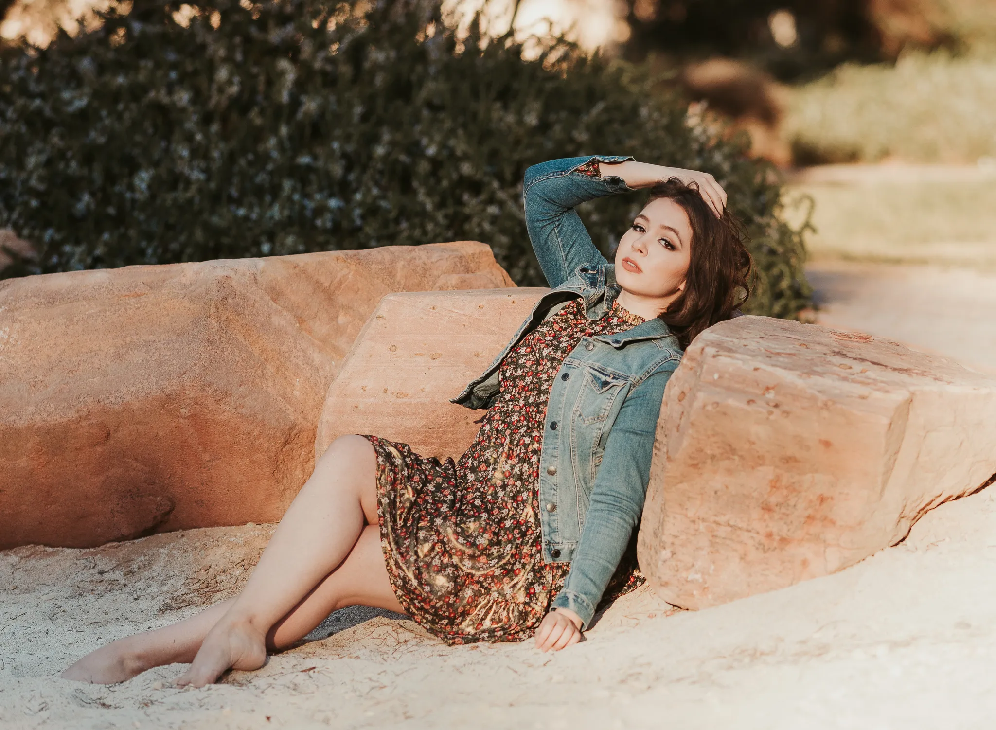 Senior in a floral dress and denim jacket reclining on red rocks at sunset