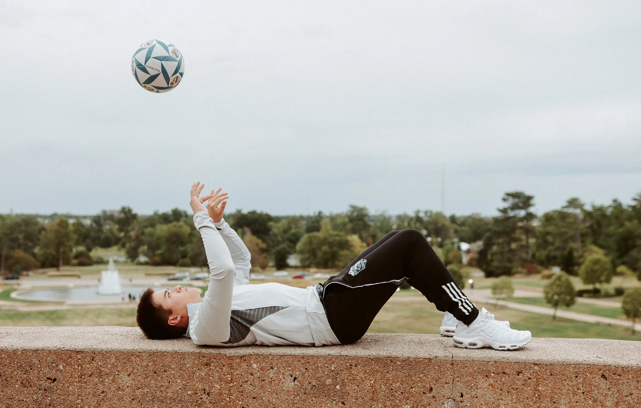 Soccer player lying on a stone ledge tossing a ball above the park fountain