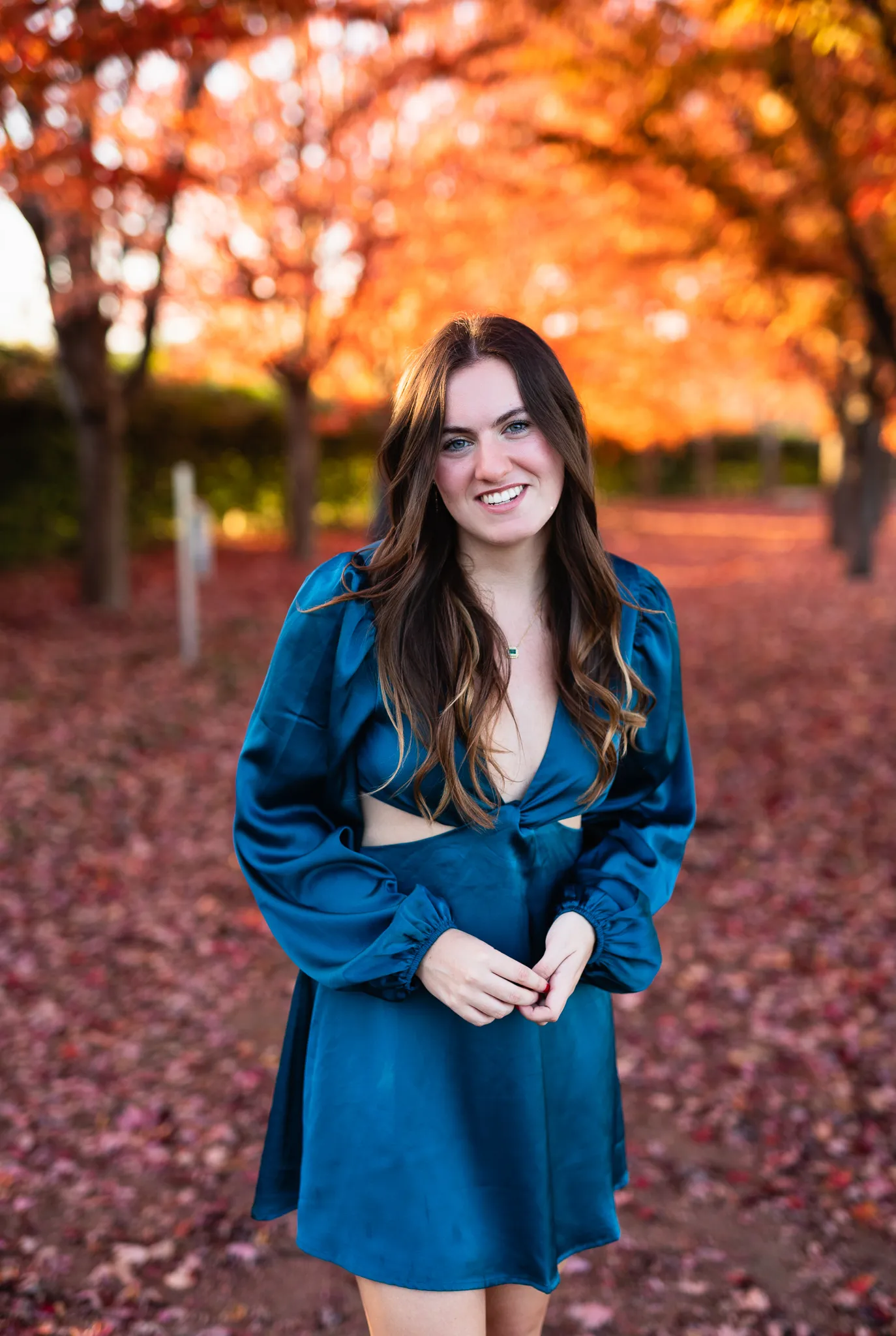Senior in a teal satin dress smiling beneath glowing autumn leaves