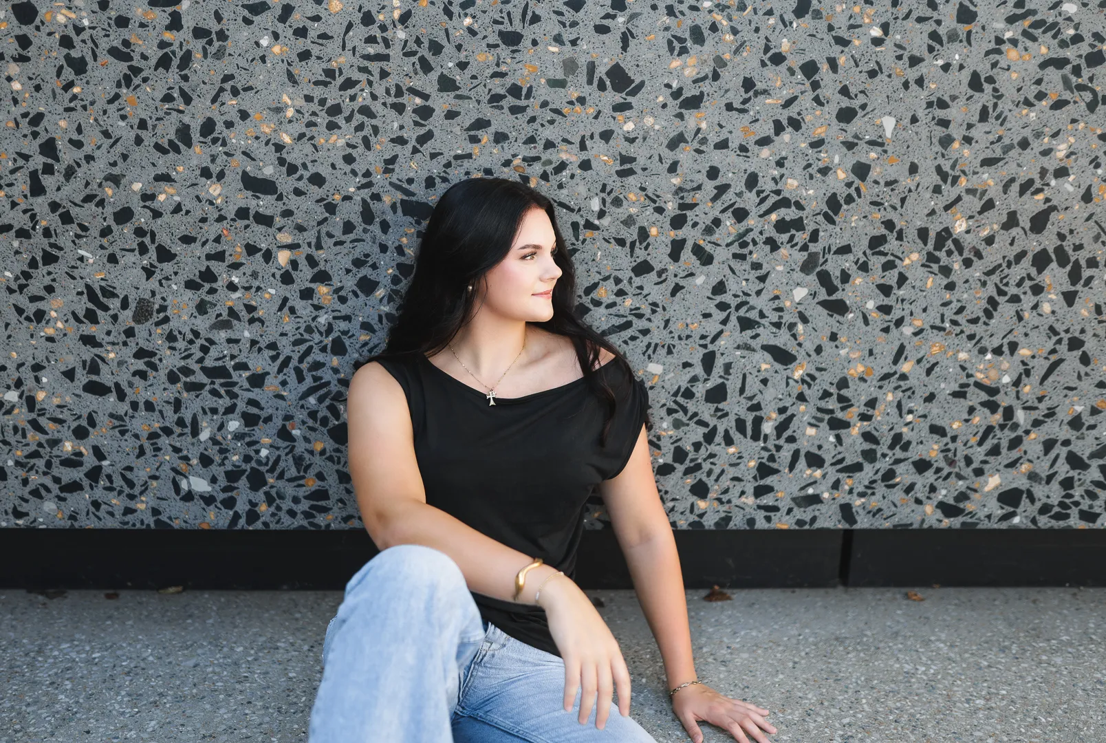 Senior in a black top and jeans seated against a terrazzo wall, looking into the light