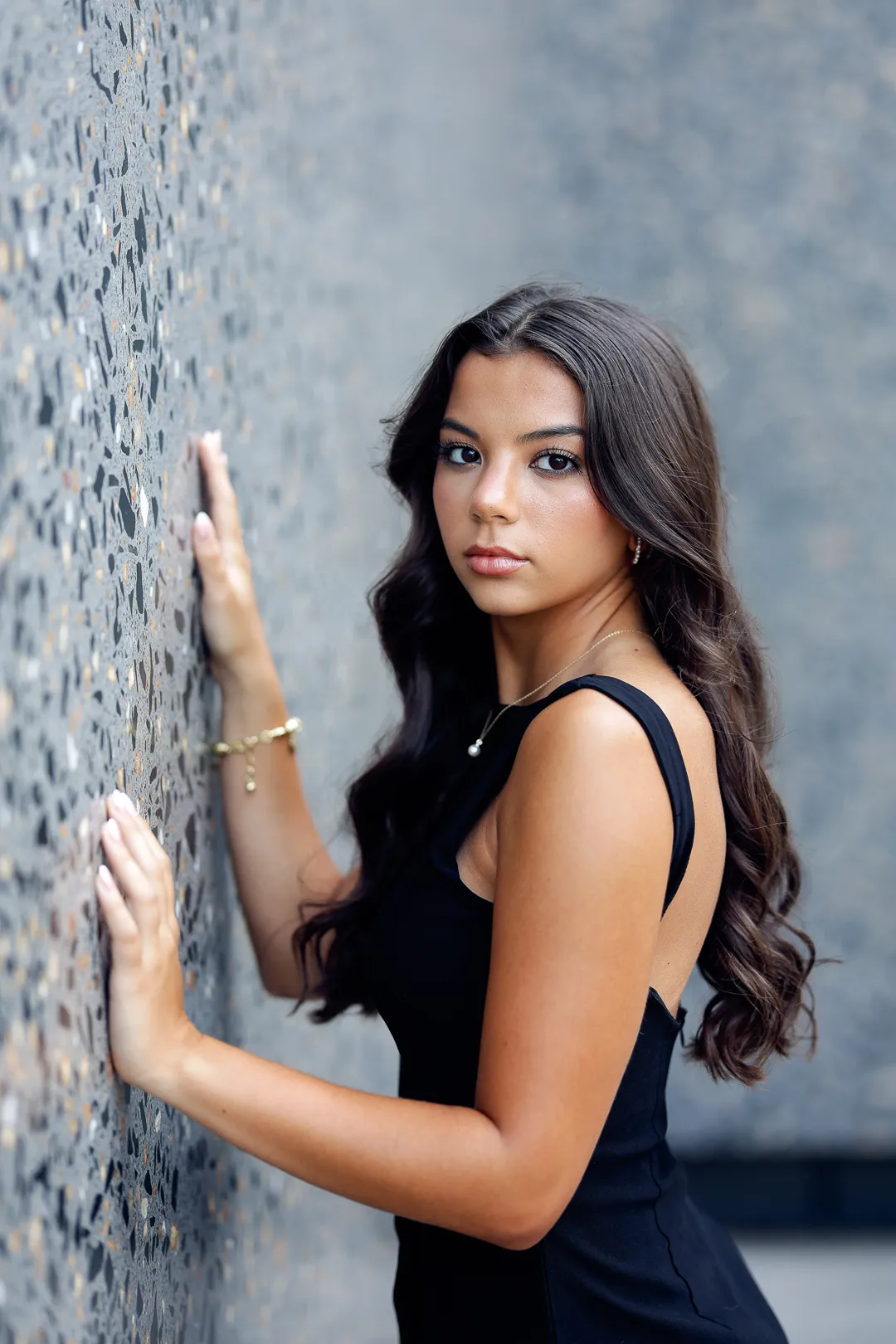 Senior in a black dress with her hands on a terrazzo wall, looking back at the camera